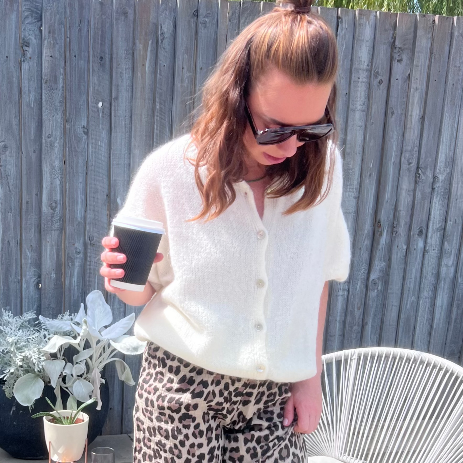 Woman standing on a patio holding a coffee cup, wearing a cream knitted cardigan and leopard print pants by penn and reid