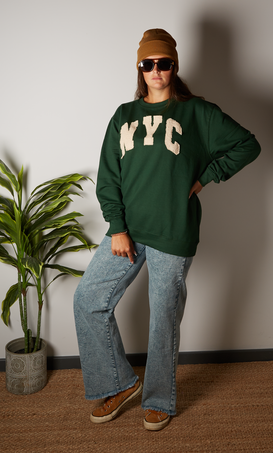 Person wearing a green 'NYC' sweatshirt, blue jeans, and brown shoes indoors.