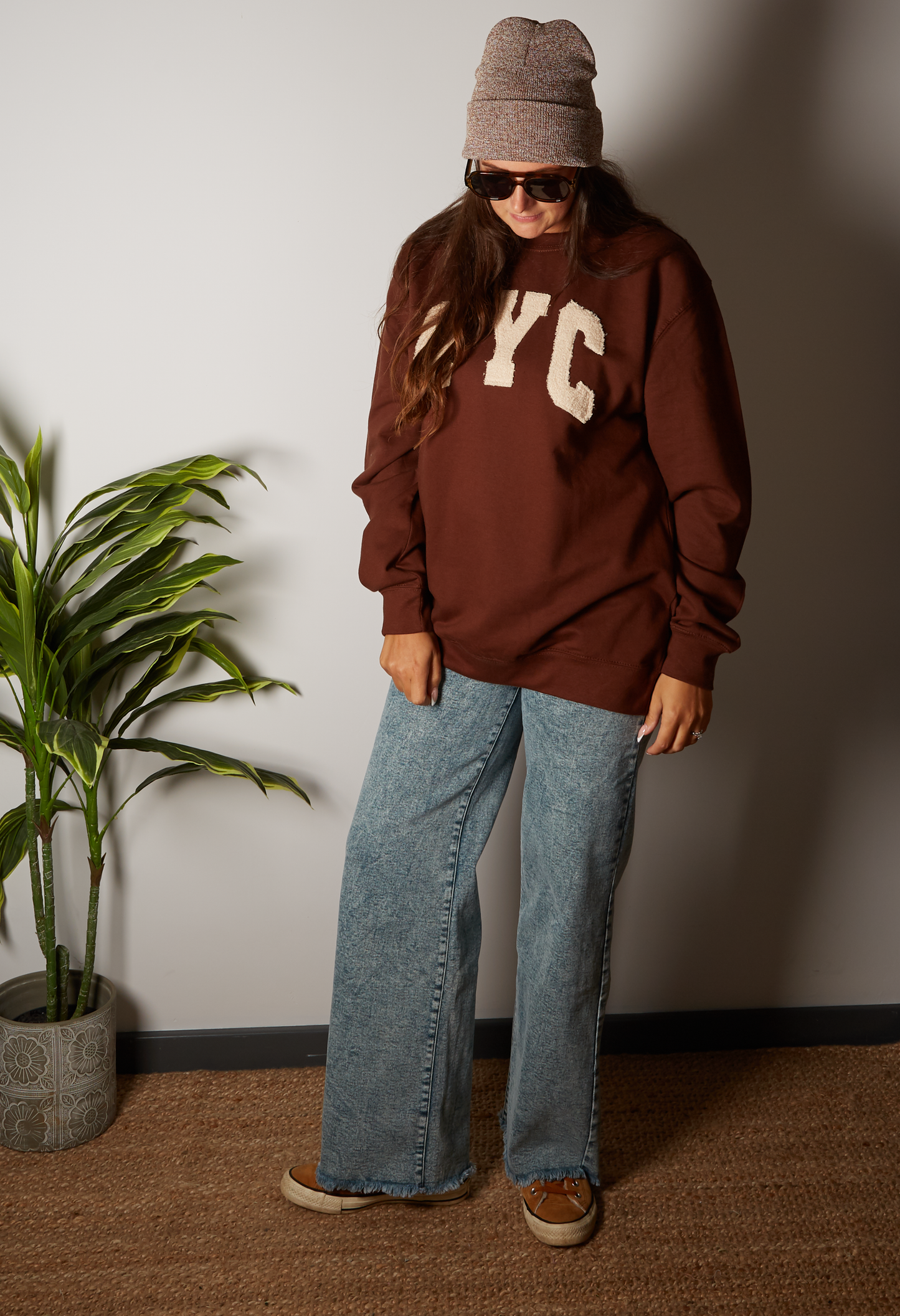 Person wearing a brown sweatshirt with 'NYC' letters, blue jeans, and a beanie standing against a plain wall.