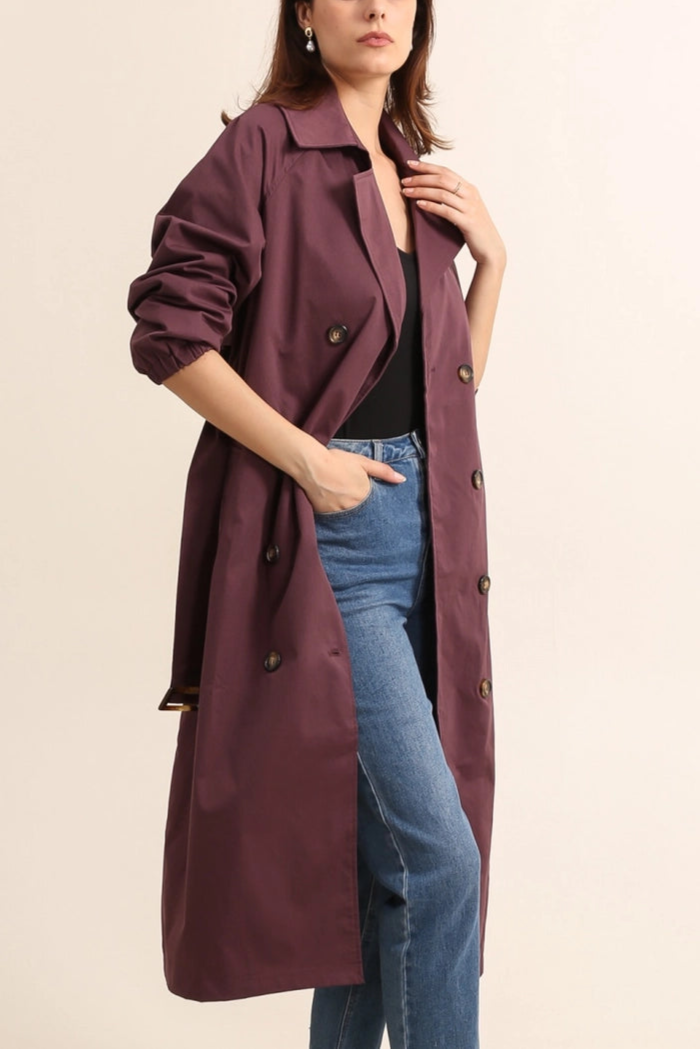 Woman wearing a becky burgundy trench coat over a black sweater and dark jeans on a plain background by penn and reid boutique leighton buzzard
