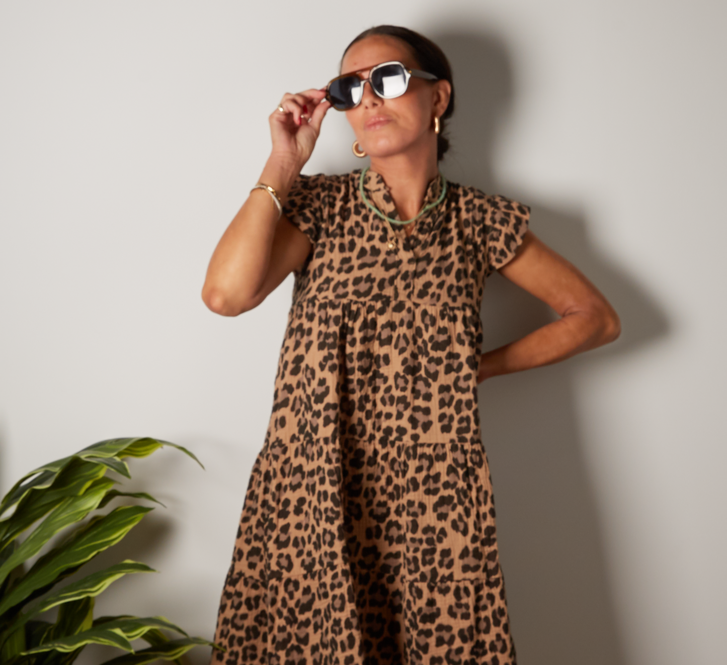 Woman wearing the harper leopard print cotton dress standing against a plain wall with a plant to her left. The dress is by Penn and Reid boutique